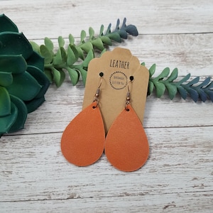 May include: A pair of teardrop-shaped leather earrings in a burnt orange color. The earrings are hanging from copper-toned hooks. The earrings are on a brown card with the text "LEATHER" and "HANDMADE JUST FOR YOU".