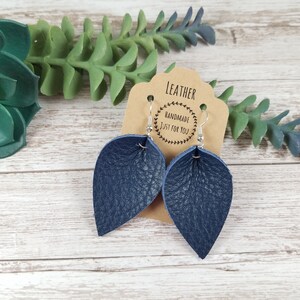 May include: A pair of dark blue leather earrings with silver hooks. The earrings are shaped like teardrops and have a textured surface. The earrings are on a brown card with the text "LEATHER HANDMADE JUST FOR YOU".