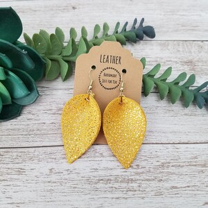 May include: A pair of teardrop-shaped earrings made of yellow textured leather with a gold glitter finish. The earrings hang from gold-toned metal hooks and are attached to a tan cardboard display card printed with the text LEATHER HANDMADE JUST FOR YOU.