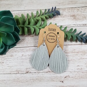 May include: A pair of light blue leather teardrop earrings with a silver hook closure. The earrings are handmade and have a woven texture. The earrings are displayed on a brown card with the text "LEATHER HANDMADE JUST FOR YOU".