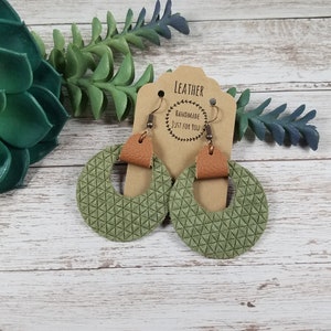 Pode incluir: Um par de brincos de couro verde com um padrão geométrico. Os brincos são feitos à mão e têm um detalhe em couro castanho. Os brincos estão em um cartão castanho com o texto "Leather Handmade Just For You".