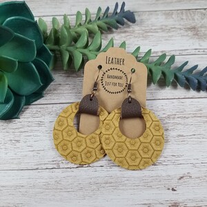 May include: A pair of mustard yellow leather earrings with a honeycomb pattern and brown leather accents. The earrings are hanging from a card that says "Leather Handmade Just For You".