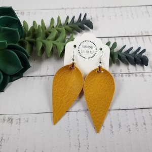 May include: A pair of mustard yellow leather earrings with a teardrop shape. The earrings are hanging from silver hooks and are on a white card with the text "Handmade Just For You".