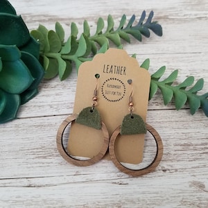 May include: A pair of handmade wooden hoop earrings with olive green suede accents. The earrings hang from copper-colored ear wires. A tan tag behind the earrings reads "LEATHER" and "HANDMADE JUST FOR YOU."