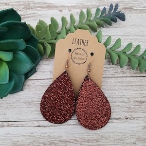Op de afbeelding: Een paar druppelvormige oorbellen met een glinsterende, roodbruine afwerking. De oorbellen hangen aan koperkleurige haken en worden gepresenteerd op een bruine kaart met de tekst "LEATHER" en "HANDMADE JUST FOR YOU". Groen gebladerte is zichtbaar op de achtergrond.