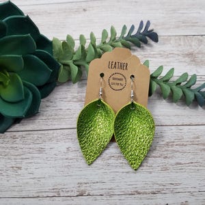May include: A pair of lime green leather earrings with a teardrop shape. The earrings are hanging from silver hooks and are on a brown card with the text "Leather Handmade Just for You".