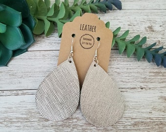 May include: A pair of silver teardrop earrings with a textured, woven appearance. The earrings hang from silver hooks and are displayed on a brown tag that reads "LEATHER" and "HANDMADE JUST FOR YOU". The earrings are set against a backdrop of green foliage and a white wooden surface.