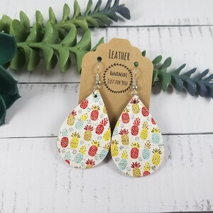 May include: A pair of teardrop-shaped leather earrings with a pineapple print. The earrings are white with red, yellow, and blue pineapples. The earrings are handmade and have silver hooks.