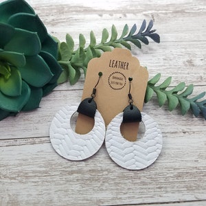 May include: White leather hoop earrings with a woven texture and black leather accents. The earrings hang from black metal hooks. A brown tag behind the earrings reads "LEATHER" and "HANDMADE JUST FOR YOU".