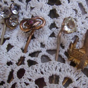 May include: A collection of antique hat pins with decorative heads. The pins feature clear rhinestones, a red gemstone, and gold-toned metal designs. One pin has a floral design, and another has a laurel wreath with the letters "GRCO".