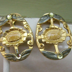 Könnte beinhalten: Ein Paar goldfarbene Ohrclips mit floralem Design und einem hellbraunen Mittelstück. Die Ohrringe haben einen schwarzen Emaille-Rand.