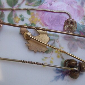 May include: A collection of antique hat pins in various designs. The pins are made of gold and silver-toned metals, with decorative elements including a knot, a floral design, and a twisted shaft. The pins are displayed on a floral patterned surface.