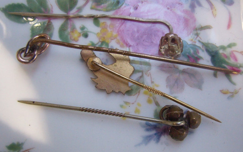 May include: A collection of antique hat pins in various designs. The pins are made of gold and silver-toned metals, with decorative elements including a knot, a floral design, and a twisted shaft. The pins are displayed on a floral patterned surface.