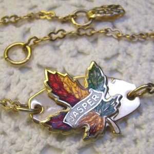 May include: A gold chain bracelet with a white enamel charm featuring a colorful maple leaf and the word "JASPER".