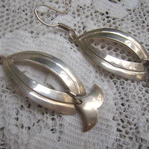 May include: A pair of silver-toned earrings. Each earring features a curved, open oval design with a stylized tail-like element at the bottom. The earrings have a hook closure and are displayed on a white lace surface.
