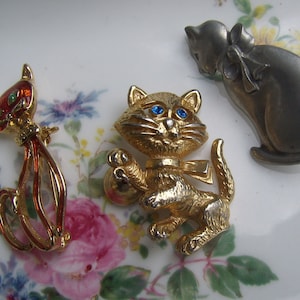 May include: Three gold tone cat brooches. One is red with green eyes, one is gold with blue eyes and a bell, and one is silver with black eyes.