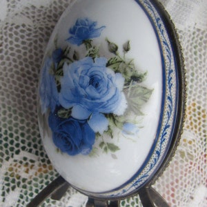 May include: A white porcelain egg-shaped trinket box with a blue and gold floral design. The box has a hinged lid and a metal base with two legs.