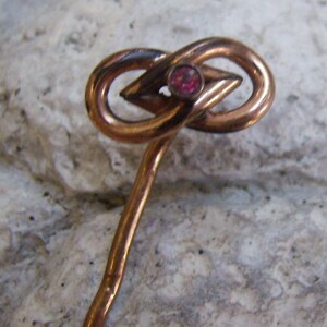 May include: A vintage copper-toned hat pin with an infinity knot design. The pin features a central diamond-shaped element and a small, round, red gemstone. The pin is designed with a long, slender stem.
