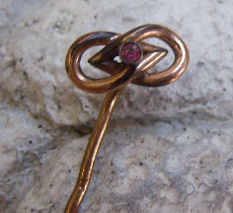 May include: A vintage copper-toned hat pin with an infinity knot design. The pin features a central diamond-shaped element and a small, round, red gemstone. The pin is designed with a long, slender stem.
