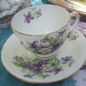 Peut inclure: Une tasse à thé et une soucoupe blanches avec une bordure dorée et un motif floral de violettes pourpres.