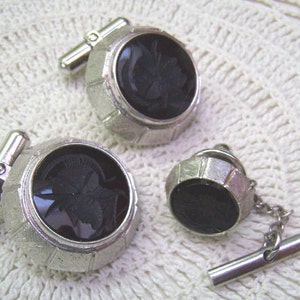 May include: A set of three silver cufflinks and a tie tack. Each piece features a black cameo with a detailed design.