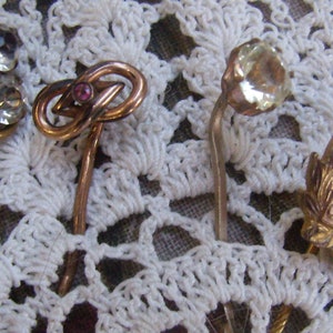 May include: A collection of vintage hat pins with decorative tops. The pins feature clear rhinestones, a knot design with a pink stone, a faceted clear stone, and a gold-toned pin with the letters "CRCO".