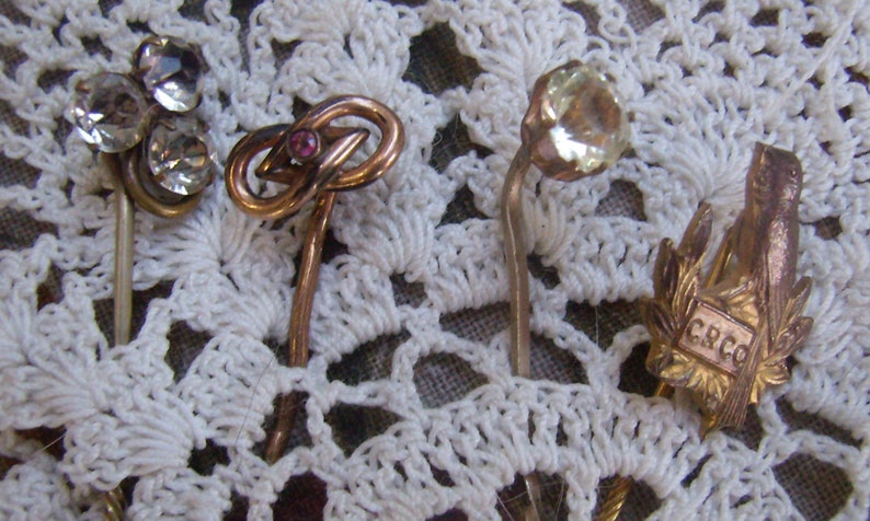 May include: A collection of vintage hat pins with decorative tops. The pins feature clear rhinestones, a knot design with a pink stone, a faceted clear stone, and a gold-toned pin with the letters "CRCO".