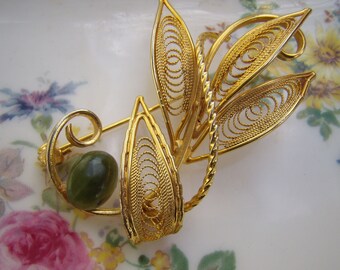 May include: A gold filigree brooch with a green stone in the center. The brooch is shaped like a leaf and has a delicate design.