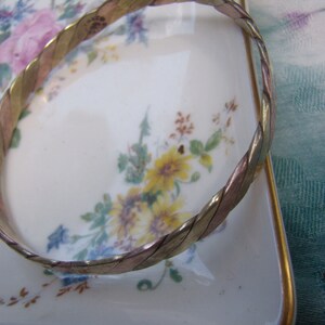 Op de afbeelding: Een gevlochten metalen armband met een gouden, zilveren en koperen afwerking. De armband ligt op een wit porseleinen bord met een bloemenmotief.