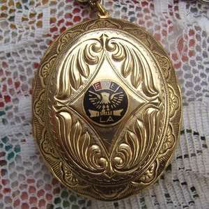 May include: Gold oval locket with an ornate design. The locket features a black enamel emblem with a white eagle, the letters "FOE" and "LA" in white, and red, white, and blue accents.