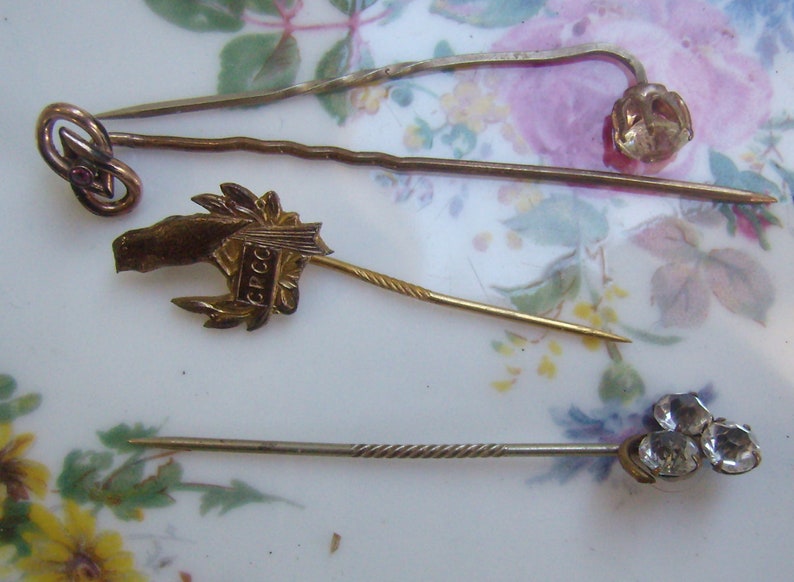 May include: A collection of antique hat pins. The pins feature various designs, including a knot with a red stone, a horse head with text, a flower, and three clear stones. The pins are gold and silver in color.
