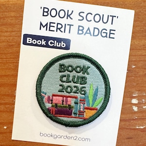 May include: A circular embroidered patch featuring a light blue background with the text 'BOOK CLUB 2026' in dark green. The design includes a stack of colorful books and a potted plant. The patch is mounted on a white card labeled 'BOOK SCOUT' MERIT BADGE.
