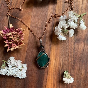 May include: A copper chain necklace with a green gemstone pendant wrapped in copper wire. The pendant is on a wooden surface with dried white flowers.
