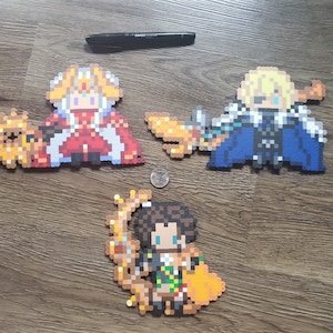 May include: Three pixel art figures made of colorful beads. The figures are of a person in a red and white robe, a person in a blue and white robe, and a person in a brown and yellow outfit. The figures are on a wooden surface.