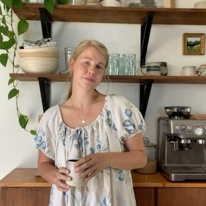 May include: A woman wearing a white dress with blue floral patterns, holding a mug. The dress has short sleeves and a round neckline. A wooden shelf with various items is visible in the background, including plants and decorative objects.