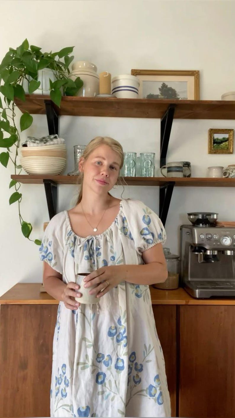 May include: A woman wearing a white dress with blue floral patterns, holding a mug. The dress has short sleeves and a round neckline. A wooden shelf with various items is visible in the background, including plants and decorative objects.