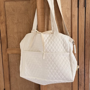 May include: A white quilted tote bag with a shoulder strap and a front pocket. The bag has a diamond pattern and a cream-colored strap. The bag is hanging on a wooden cabinet.