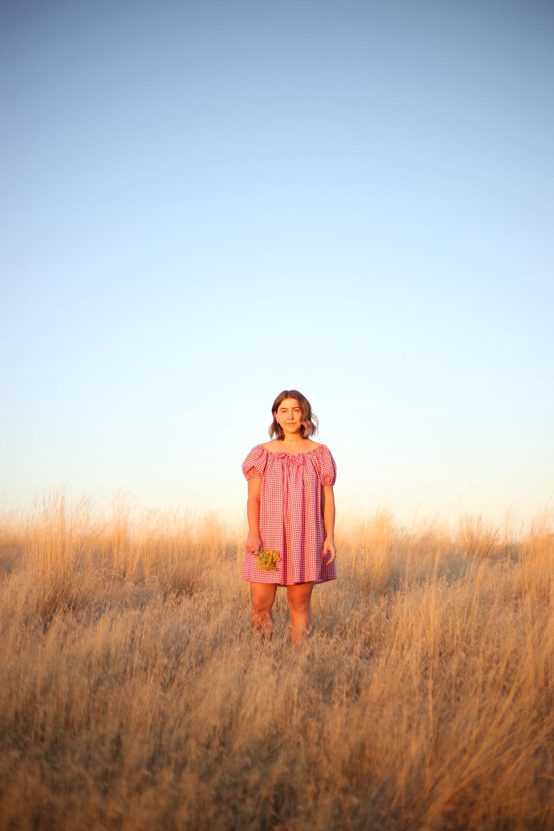 May include: A person stands in a field of tall, dry grass, wearing a short, red and white gingham dress. The dress has short sleeves and a gathered neckline. The person holds a small bouquet of yellow flowers. The sky is a gradient of blue and orange.
