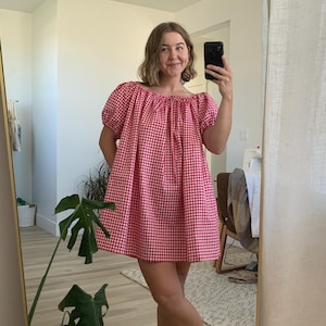 May include: A red and white gingham print dress with an off-the-shoulder neckline and short, puffed sleeves. The dress is knee-length and appears to be made of a lightweight fabric. The image is taken in front of a mirror.
