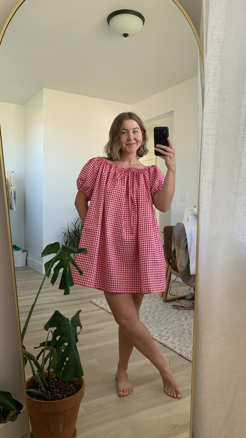 May include: A red and white gingham print dress with an off-the-shoulder neckline and short, puffed sleeves. The dress is knee-length and appears to be made of a lightweight fabric. The image is taken in front of a mirror.