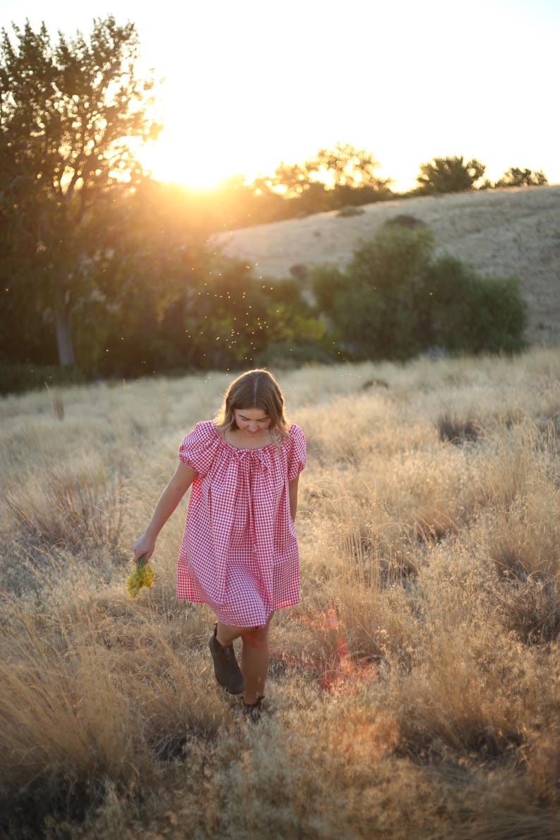 May include: A person wearing a red and white gingham dress walks through a field of tall, dry grass. The dress has short sleeves and a scoop neckline. The person is holding a small bouquet of yellow flowers. The sun is setting in the background.