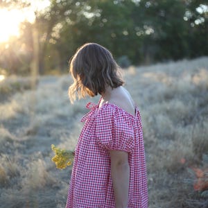 May include: A person wearing a red and white gingham dress with off-the-shoulder sleeves and tied bows. The dress is loose-fitting and the person is holding a small bouquet of yellow flowers. The background is a field of dry grass with sunlight.