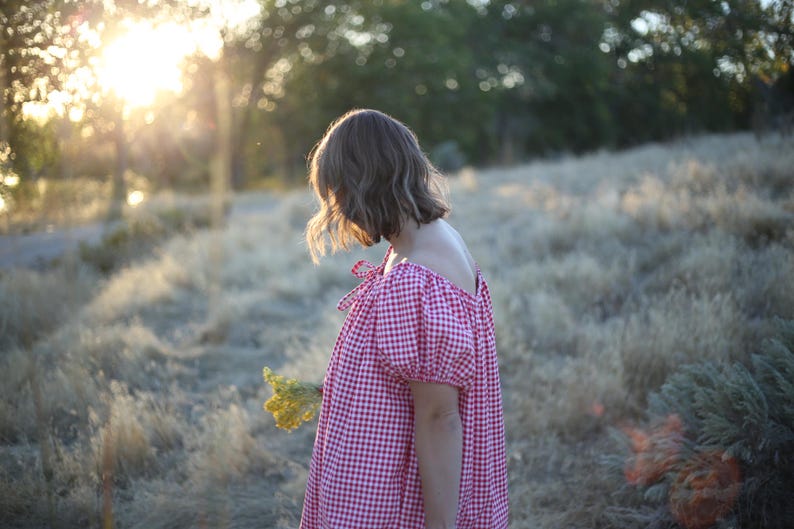 May include: A person wearing a red and white gingham dress with off-the-shoulder sleeves and tied bows. The dress is loose-fitting and the person is holding a small bouquet of yellow flowers. The background is a field of dry grass with sunlight.