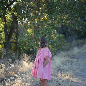 May include: A person wearing a red and white gingham dress with short, puffed sleeves. The dress is knee-length and has pockets. The background features a dirt road, dry grass, and trees with green and yellow leaves.