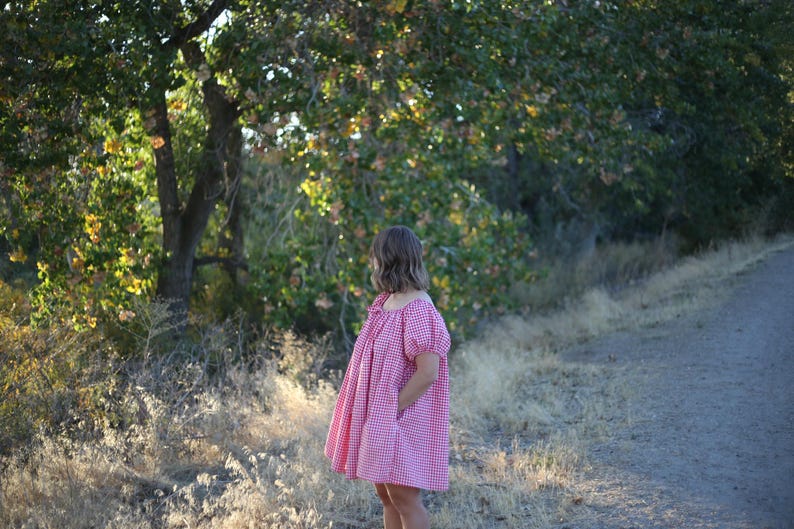 May include: A person wearing a red and white gingham dress with short, puffed sleeves. The dress is knee-length and has pockets. The background features a dirt road, dry grass, and trees with green and yellow leaves.