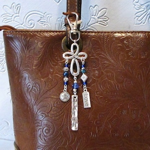 May include: A brown leather purse with a silver charm attached to the zipper. The charm features a cross with blue and silver beads and three silver tags that read "Hope", "Faith", and "Love".