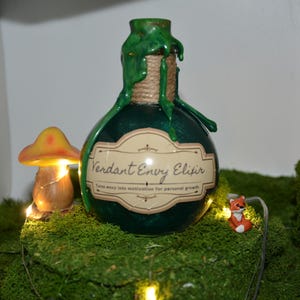 May include: A dark green bottle labelled "Verdant Envy Elixir" rests on green moss, with a mushroom and fox figurine. The bottle has green drippings and is tied with twine. The scene is lit with small fairy lights.