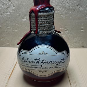 Może przedstawiać: Czarna szklana butelka z czerwoną pieczęcią woskową i etykietą z napisem "Rebirth Draught. Encourages boldness and joyful spontaneity."