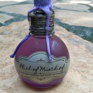 May include: A small glass bottle with a purple liquid and a label that reads "Phial of Mischief" and "Encourages boldness and joyful spontaneity." The bottle is topped with a purple wax seal and tied with twine.
