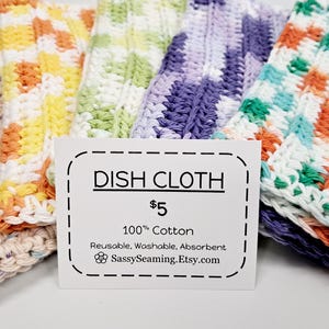 May include: A collection of colorful, handmade dish cloths in various patterns and colors, including cream, yellow, orange, green, purple, and teal. The cloths are made of 100% cotton and are reusable, washable, and absorbent. A sign reads "Dish Cloth".